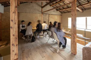 A casual indoor gathering area with wooden tables and chairs, natural light, and people socializing and enjoying meals