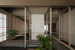 A calm and inviting indoor space with a large white wall, wooden structure, and several potted plants, including one near the center and another near the right side, with a chair in the background