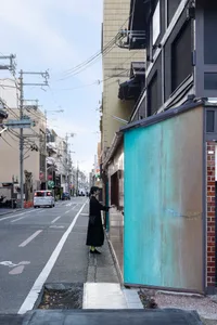 一位女子站在大樓旁的行人道上，看著前方某物，附近停放數輛汽車與機車，處於熱鬧的城市環境中