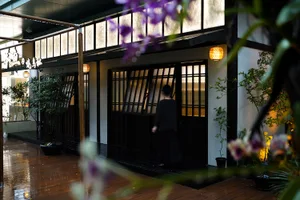An Asian woman walks through a building with sliding doors, surrounded by oriental design, potted plants, and a wooden floor