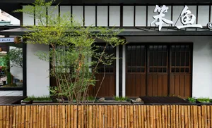 Asian-style building with wooden walls, bamboo fencing, a balcony, and a doorway leading to a patio area, surrounded by two potted plants