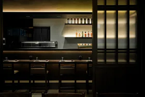 A cozy bar area with a long wooden counter and chairs, featuring bottles and a welcoming atmosphere