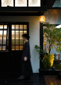 A woman in a black dress walks down a hallway with a white wall, a potted plant, two vases, and a bowl
