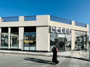 一位女子走在建築物前方的休閒街景，周圍停有汽車、戶外家具與餐桌，背景為輕鬆的都市景觀