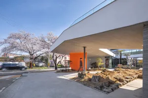 A modern building with a large glass wall extending over a walkway, surrounded by trees and rocks, with people walking around outside