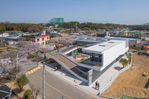 A modern building with a white exterior and a large glass wall, featuring a bridge connecting two parts and people walking around the area
