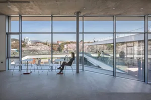 A woman sits in a chair near a window in a room with multiple chairs and dining tables, surrounded by concrete walls and glass surfaces, with a calm and relaxed atmosphere