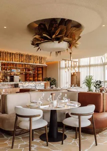 Modern restaurant interior with wooden elements, white and brown tones, and potted plants, featuring tables, chairs, and wine glasses