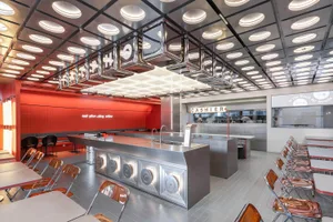 Busy commercial kitchen with a large countertop island surrounded by chairs and several people working or preparing food