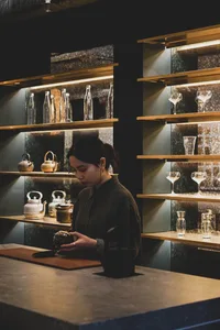 A woman stands in front of a wooden counter filled with wine glasses, cups, and bottles, surrounded by a social gathering atmosphere