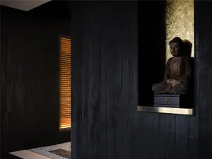 A dark room with a wooden Buddha statue on a pedestal, two windows, and a solemn atmosphere