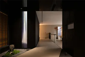 A modern living space with a black wall featuring a waterfall, a dining table, and a chair, with a glass door and a second door, set in a stylish and minimalist atmosphere