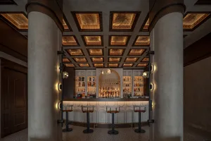 A cozy bar area with a wooden counter, multiple chairs, and paintings on the walls, featuring a selection of bottles and comfortable seating for patrons