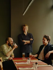 Three people sitting around a wooden dining table in a relaxed restaurant setting, enjoying beer and conversation