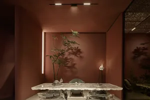 A dining area with a marble table surrounded by chairs, set against a warm red-hued backdrop with natural light pouring in