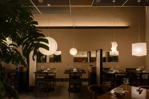 A warm and inviting dining area with multiple tables, chairs, and potted plants, featuring a mix of styles and well-designed lighting