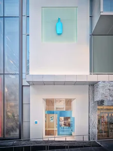 A museum interior featuring a large glass wall showcasing a blue-hued wooden sculpture, with two bottles in the scene, creating an artistic and creative atmosphere