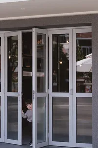 A young child stands in front of a glass door with a silver frame, peeking through the sliding doors with a curious expression