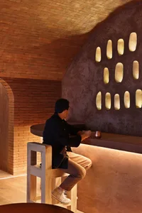 A man sits at a wooden bar in a cozy cafe with a sink and two chairs, enjoying a cup of coffee in a relaxed atmosphere