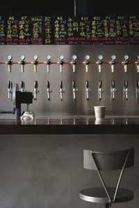 Industrial-style bar area with a long counter featuring 14 beer taps and two cups, set in a busy restaurant or bar atmosphere