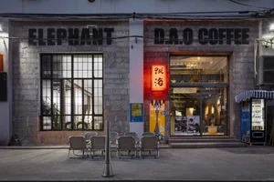 A brick coffee shop with a red neon sign, outdoor seating, and potted plants, set in an urban street scene