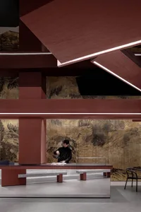 A man prepares food at a wooden counter in an Asian restaurant with a red wall and two chairs