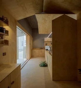 Open modern kitchen and living space with wooden walls and flooring, featuring a refrigerator, microwave, and oven, with potted plants and a vase adding warmth and coziness