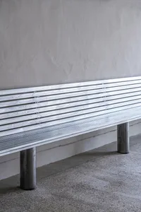 A metal bench in a minimalist style sits in front of a wall, surrounded by a bright and welcoming atmosphere, suggesting a daytime photo shoot on a sunny day