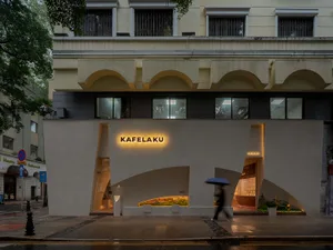 A modern city street scene with a large white building featuring a prominent archway, surrounded by urban elements and a potted plant