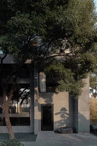 A gray building with a tree in front, casting a shadow on its surface, and people present in the scene