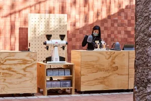 A woman stands behind the counter in a cozy coffee shop with two wooden tables and various cups and bottles scattered around the area