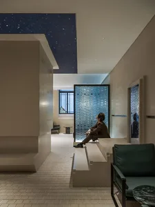 A modern living area with a man sitting on a bench in front of a window, surrounded by two chairs and concrete walls that allow natural light to enter