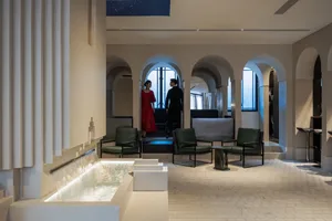 Spacious and luxurious interior design with high ceilings, well-lit and featuring chairs, a bench, and two people standing in the room
