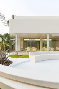 Open modern living area with white walls, floors, and chairs, and two dining tables in a bright and airy space