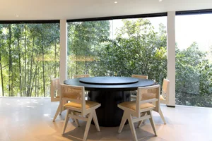 Dining area with a round wooden table and modern chairs, surrounded by a large window with natural light
