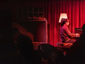 A woman plays the piano in a dimly lit room with a couch and two people present