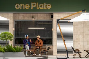 Two men sit in a modern cafe with a storefront window, surrounded by wood and glass elements, potted plants, and a casual atmosphere