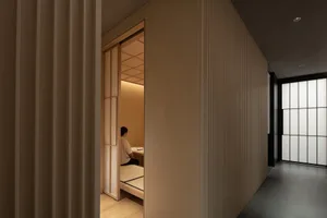 A small bedroom or living area with wooden floors and walls, featuring a person sitting on a bed and another near the doorway