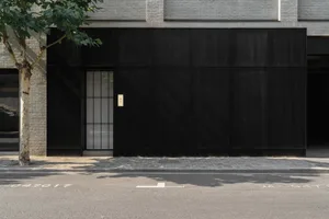 Exterior view of a brick building with a black doorway and glass window above, set against a neutral background