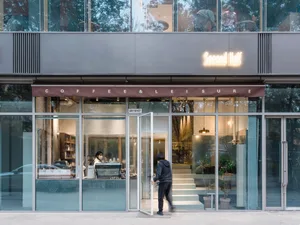 A spacious coffee shop with a glass door entrance, couches, chairs, potted plants, and scattered books, creating a cozy atmosphere for reading and relaxation