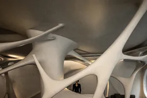 A woman stands in front of a large white concrete sculpture resembling a spiral staircase in a modern and minimalist lobby or public space