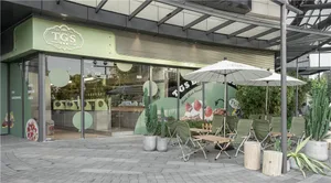 An outdoor cafe with chairs and tables in front of a green building, shaded by umbrellas
