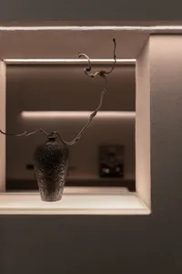 A dimly lit room with a metal vase on a table against a white wall, minimalist composition