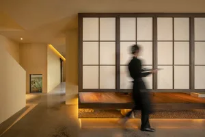 A woman walks through a dimly lit hallway with a wooden bench and a door and window on either side