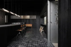 Dark and moody restaurant kitchen with black walls and floors, featuring empty chairs and a minimalist layout