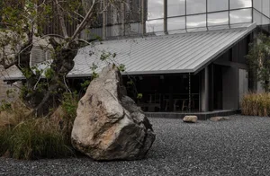 A building with a gray roof and a large rock in front, surrounded by outdoor seating and natural surroundings