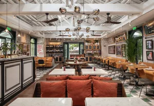 A restaurant interior with multiple tables, couches, and paintings, illuminated by hanging lights, creating a warm and inviting atmosphere