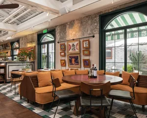 Cozy restaurant dining area with multiple couches and chairs around a large round table, wine bottles on the table, and a warm atmosphere