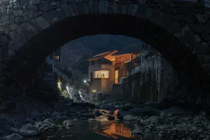 A stone house near a river with a peaceful atmosphere, two people standing outside, and a serene environment
