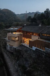 Asian-style house with a wooden exterior, situated on a hillside overlooking a river, with a large open area leading to a patio and several windows allowing natural light